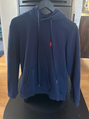 Marinblå hoodie från Polo Ralph Lauren - Snygg marinblå hoodie från Polo Ralph Lauren med klassisk röd logga på bröstet. Modellen har huva med dragsko och en stor magficka framtill. Tillverkad i mjukt bomullsmaterial och har ribbade muddar vid ärmslut och nederkant. Perfekt för en avslappnad och stilren look.