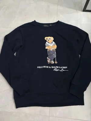 Svart Polo Bear sweatshirt Ralph Lauren - Säljer en svart sweatshirt från Ralph Lauren med den ikoniska Polo Bear-trycket framtill. Tröjan har rund hals, långa ärmar och ribbade muddar. Perfekt för dig som gillar klassisk streetwear med en twist. Materialet är mjukt och bekvämt, troligen bomullsblandning.
