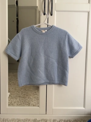 Ljusblå stickad t-shirt Gina Tricot - Supersöt ljusblå stickad t-shirt från Gina Tricot. Tröjan har korta ärmar, rund halsringning med ribbad kant och är gjord i ett mjukt, fluffigt material. Perfekt för lager på lager eller som ett statement-plagg till jeans eller kjol.