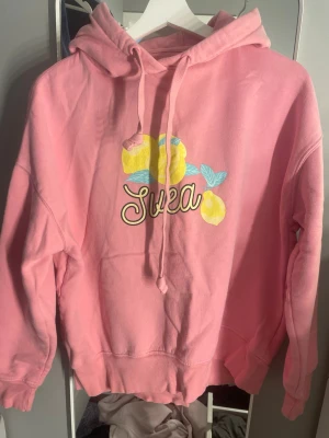 Rosa hoodie med citroner från Svea - Säljer en rosa hoodie från Svea med tryck av gula citroner och turkosa blad på bröstet samt Svea-loggan i vitt. Hoodien har huva med dragsko och ribbade muddar vid ärmar och nederkant. Perfekt för dig som gillar färgglada och lekfulla plagg.