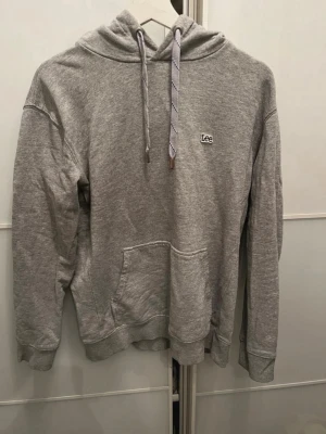 Grå hoodie från Lee  - Säljer en klassisk grå hoodie från Lee med broderad logga på bröstet. Hoodien har huva med dragsko, magficka och ribbade muddar vid ärmar och nederkant. Tillverkad i mjukt sweatshirtmaterial, perfekt för en avslappnad stil.