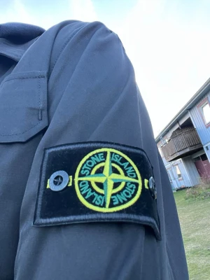 Svart jacka från Stone Island - Säljer en svart jacka från Stone Island med klassisk logotyp på ärmen. Jackan har dragkedja framtill, bröstficka med lock och en stilren design. Perfekt för dig som gillar streetwear och vill ha något snyggt och funktionellt. Helt ny, använd 1 gång 