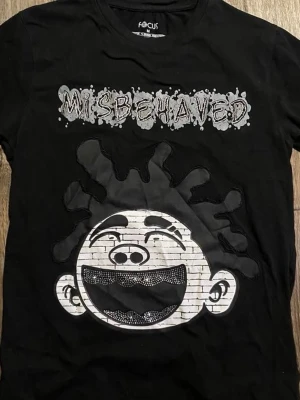 Svart t-shirt med tryck från Focus - Cool svart t-shirt från Focus med stort grafiskt tryck av ett leende ansikte och texten 'MISBEHAVED' i vitt och grått. T-shirten har rund hals och korta ärmar, perfekt för en avslappnad streetwear-look.