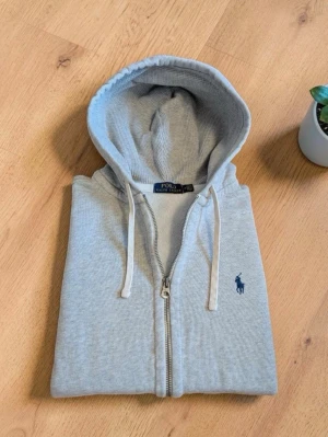 Grå hoodie från Polo Ralph Lauren - Snygg ljusgrå hoodie med dragkedja från Polo Ralph Lauren. Klassisk design med huva och vita snören, samt den ikoniska blå loggan broderad på bröstet. Tillverkad i mjuk bomullsblandning och har kängurufickor framtill. Perfekt för en chill och stilren look.