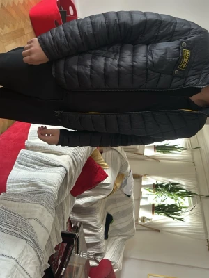 Svart Barbour quiltad jacka - Säljer en svart quiltad jacka från Barbour med gul insida och dragkedja. Jackan har en bröstficka med Barbour-logga och är tillverkad i ett lätt, vadderat material. Perfekt för dig som vill ha en stilren och bekväm jacka.