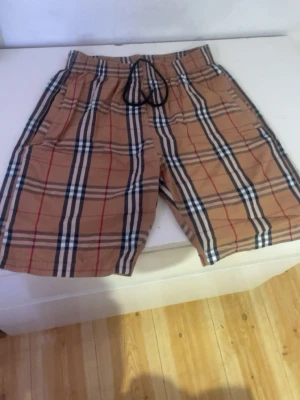  shorts från Burberry - Säljer ett par klassiska beige shorts från Burberry med det ikoniska rutiga mönstret i svart, vitt och rött. Shortsen har elastisk midja med snörning och sidofickor. Perfekta för en trendig och avslappnad stil.