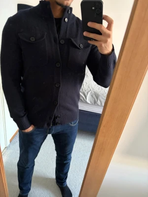 Stickad cardigan - Stilig mörkblå stickad cardigan från Less Worries i storlek S med hög krage och knäppning framtill. Koftan har två bröstfickor med lock och ribbade muddar vid ärmslut och nederkant samt extra knappar som följer med. Perfekt nu till våren! Inga defekter! Bara att kontakta vid funderingar!🌟