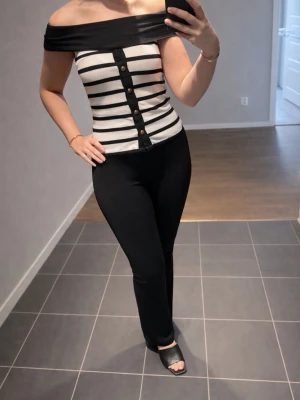 Randig offshoulder topp med knappar - Snygg offshoulder topp med svarta och vita ränder och dekorativa knappar framtill. Toppen har en bred svart kant upptill och är figurnära med axelbandslös design. Perfekt för dig som vill sticka ut lite extra.