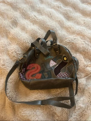 Grå handväska från Desigual  - Unik grå handväska från Desigual med färgglada detaljer på framsidan. Väskan har både handtag och justerbar axelrem i samma material. Stängs med dragkedja och har detaljer i metall. 