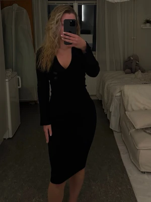 Svart långärmad klänning med v-ringning - En stilren svart klänning med lång ärm och v-ringad hals. Klänningen har en kroppsnära passform och slutar strax under knäet. Perfekt för dig som gillar en klassisk och elegant look.