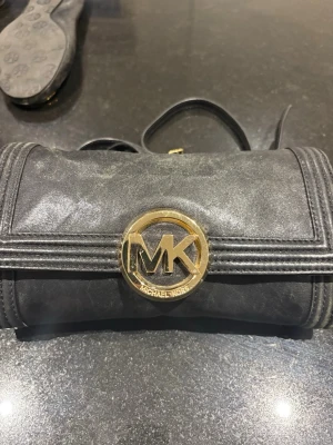 Svart kuvertväska Michael Kors - Snygg svart kuvertväska från Michael Kors med stort guldfärgat MK-emblem framtill. Väskan är i läderimitation och har ett stilrent, klassiskt utseende med magnetlås och innerfack med dragkedja. Perfekt för att lyfta din outfit.