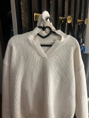 Vit stickad tröja med v-ringning - Mysig vit stickad tröja med v-ringning och ribbad struktur. Tröjan har långa ärmar och en avslappnad passform. Materialet ser ut att vara mjukt och stretchigt, vilket gör den extra bekväm.