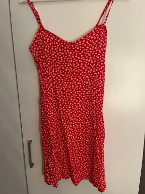 Röd blommig sommarklänning - Söt röd kortklänning med vita blommor och smala axelband. Klänningen har en lätt utsvängd passform och är perfekt för varma dagar. Materialet känns mjukt och luftigt, vilket gör den skön att bära. Ingen synlig logga eller märke. Köpt från newyorker. 