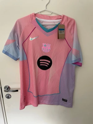 Barca x Nike rosa matchtröja - Säljer en FC Barcelona matchtröja från Nike i rosa och ljusblått med klubbmärke och Spotify-logga på bröstet. Tröjan har korta ärmar, V-ringning med turkos kant och är tillverkad i lätt, ventilerande funktionsmaterial. Perfekt för fotbollsträning eller support. Tröjan är helt ny och aldrig använd😃