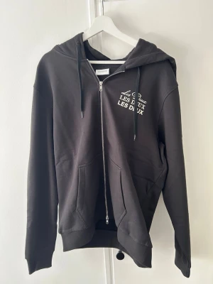 Svart zip-hoodie från Les Deux - Svart hoodie med dragkedja från Les Deux. Tryck i vitt på bröstet, två fickor framtill och justerbar huva med snören. Klassisk och stilren modell i mjukt material, perfekt för en avslappnad look.