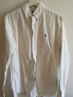 Vit skjorta från Polo Ralph Lauren - Klassisk vit skjorta från Polo Ralph Lauren i slim fit-modell. Skjortan har långa ärmar, knappar framtill och den ikoniska broderade loggan på bröstet. Tillverkad i mjuk bomull och passar perfekt till en stilren look.