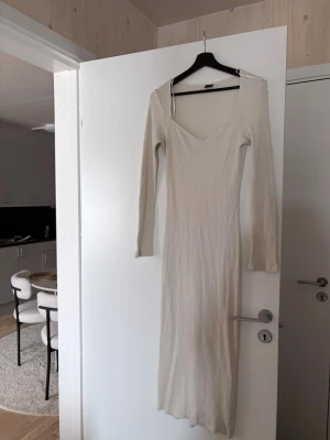 Lång ribbad klänning i beige - Långärmad, ribbad klänning i beige med v-ringning och figurnära passform. Klänningen är hellång och har en stilren, enkel design som passar perfekt för en trendig look.