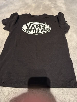 Svart Vans t-shirt med tryck - Säljer en svart t-shirt från Vans med det klassiska 'Off The Wall'-trycket i vitt på bröstet. T-shirten har rund hals och korta ärmar. Perfekt för dig som gillar streetwear och vill ha en enkel men snygg look.
