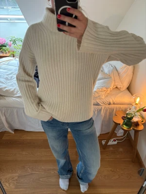 Ribbstickad polotröja i beige - Mysig ribbstickad polotröja i beige med hög krage och långa ärmar. Perfekt för kyliga dagar och enkel att matcha med jeans eller kjol. Tröjan har en loose passform och är tillverkad i ett mjukt stickat material.