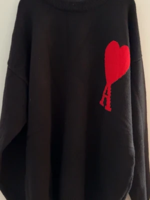 Svart stickad tröja med rött hjärta Ami - Säljer en svart stickad tröja från AMI Paris med deras ikoniska röda hjärta och bokstaven A broderat på bröstet. Tröjan har rund halsringning, långa ärmar och ribbade muddar. Perfekt för dig som gillar streetstyle och vill ha något trendigt. Helt ny