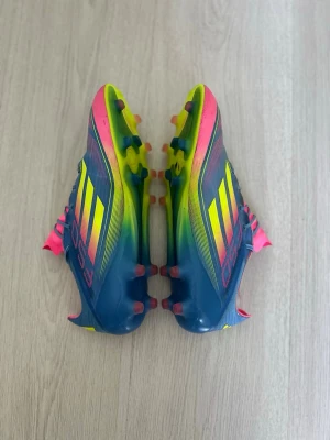 Adidas F50 Elite - Säljer ett par Adidas F50 elite fotbollsskor i blått med neon-gula och rosa detaljer. Skorna har klassisk snörning, låg profil och mönstrad sula för bra grepp på planen. Tillverkade i syntetmaterial för lätt känsla och snabbhet. Behövde klippa toppen av skosnören pga den gick sönder