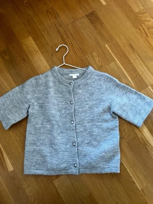 Grå kortärmad kofta från Ginatricot - Jättefin grå stickad tröja från Ginatricot med korta ärmar. Koftan har pärlemorknappar framtill! 💕☀️I storlek 170