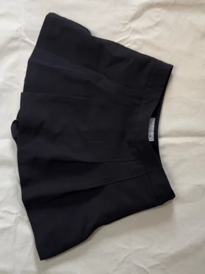 Svart skort med hög midja - Stilren svart skort med hög midja och diskreta sömdetaljer framtill. Skorten har en klassisk A-linjeform och är tillverkad i ett mjukt, vävt tyg som ger en snygg siluett.