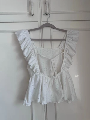 Vit volangblus med broderie anglaise - Superfin vit topp med volangärmar och broderie anglaise-mönster. Toppen har en söt knytning i ryggen och en lätt utsvängd peplumform. Perfekt för sommaren och ger en nice vibe till din outfit.