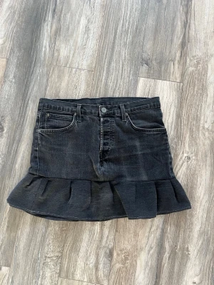 Svart jeanskjol med volangkant - Cool svart jeanskjol med volangkant nertill och klassiska fickor fram och bak. Kjolen har en kort modell och stängs med knapp och dragkedja framtill. Perfekt för dig som gillar en edgy och trendig stil.