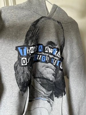 Grå hoodie från Trapstar med tryck - Säljer en grå hoodie från Trapstar med stor grafisk print framtill av Benjamin Franklin och texten 'That's an awful lot of cough syrup'. Hoodien har huva och är gjord i mjukt bomullsmaterial. Perfekt för dig som gillar streetwear och unika prints.
