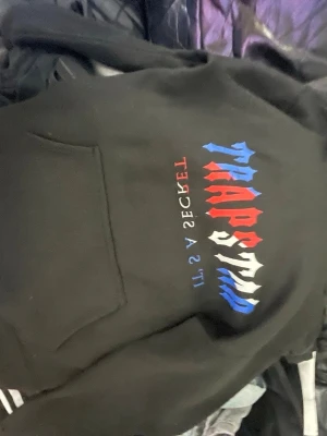 Svart Trapstar hoodie med tryck - Säljer en svart hoodie från Trapstar med färgglatt tryck i blått, rött och vitt på bröstet. Hoodien har huva och en stor magficka framtill. Perfekt för dig som gillar streetwear och vill sticka ut med en snygg logga.