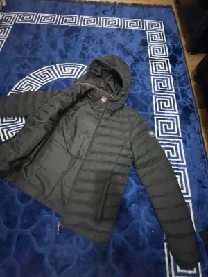 Svart pufferjacka med huva - Snygg svart pufferjacka från Stadium med quiltad design och huva med dragsko. Jackan har en diskret logga på ärmen och dragkedja framtill. Perfekt för kalla vinterdagar och riktigt skön att bära.