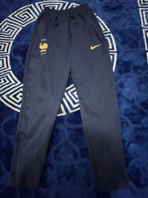 Frankrike Nike träningsbyxor marinblå - Mörkblå träningsbyxor från Nike med Frankrikes landslagslogga i guld på ena benet och Nike-logga i guld på andra. Byxorna har smal passform och elastisk midja, perfekta för sport eller chill. Materialet är troligtvis polyester för bra rörelsefrihet.