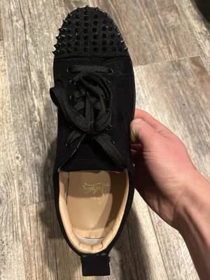 Svarta Louboutin sneakers med nitar - Säljer ett par svarta sneakers från Christian Louboutin med mattsvarta nitar på tån och klassisk röd sula. Skorna är i mocka och har svarta skosnören samt beige innersula med logga. Riktigt statement-piece för dig som gillar edgy stil.