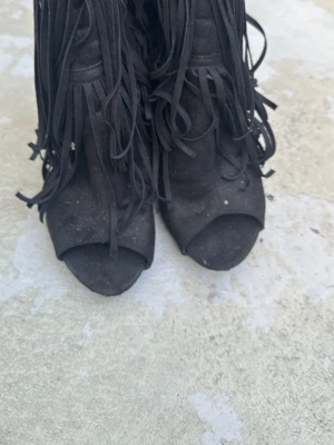 Svarta stövletter med fransar - Säljer ett par svarta stövletter med öppen tå och långa fransar i mockaimitation. Snygg detalj med guldfärgad dekoration baktill. Perfekta för dig som vill sticka ut med din stil.