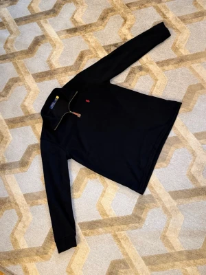 Svart stickad tröja från Polo Ralph Lauren - Svart stickad tröja med halv dragkedja från Polo Ralph Lauren. Tröjan har lång ärm och klassisk krage, med en liten röd logotyp broderad på bröstet. Perfekt för att hålla sig varm och snygg under kyliga dagar.