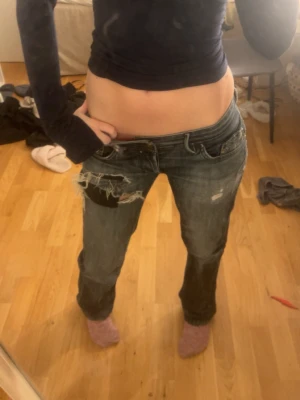Blå slitna low waist jeans - Säljer ett par blå low waist jeans med slitningar och hål på vänster lår. Jeansen har raka ben och klassisk femficksmodell. Perfekta för en avslappnad och trendig look.