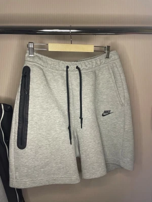 Grå Nike shorts med svart dragkedja - Snygga grå shorts från Nike med svart logga på vänster ben och en lång svart dragkedja på höger sida. Shortsens material är mjukt och bekvämt, perfekt för sport eller chill. De har elastisk midja med svarta snören för justering.
