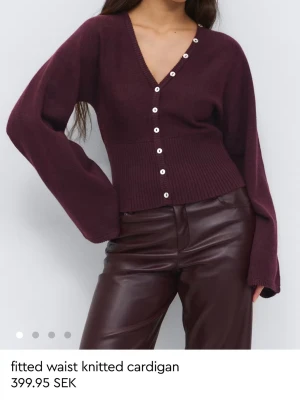 Vinröd ribbad kofta med knappar - Vinröd stickad kofta från GINATRICOT med v-ringning och vita knappar längs hela framsidan. Koftan har ribbad midja som ger en snygg passform och långa ärmar med lite lösare stil. Perfekt för att skapa en trendig look.
