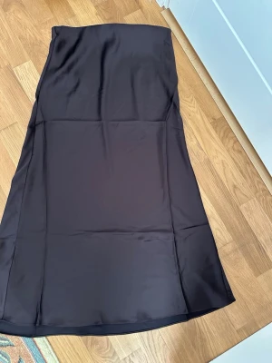 Mörkbrun långkjol i satin, storlek M - Stilren svart långkjol i mjukt satinliknande material. Kjolen har en enkel, rak passform och faller snyggt längs benen. Perfekt för dig som gillar minimalistisk och elegant stil. Mått i midjan från den ena änden till den andra änden cirka 41 cm. 