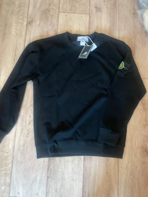 Svart sweatshirt från Stone Island - Säljer en svart sweatshirt från Stone Island med klassisk rund hals och ribbade muddar. Tröjan har den ikoniska Stone Island-loggan på vänster ärm och är tillverkad i mjuk bomull. Perfekt för en clean och stilren look.
