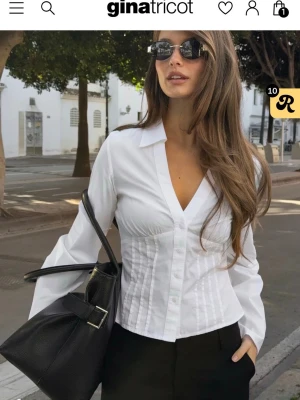 Vit figursydd skjorta från Gina Tricot - Snygg vit skjorta från Gina Tricot med figurnära passform och markerad midja. Skjortan har v-ringning, klassisk krage och knappar framtill. Långa ärmar och lätt utsvängd nederkant. Perfekt att styla med både jeans och kostymbyxor. I storlek 34/Xs. Endast använd en gång. Helt slutsåld på hemsidan💕💕