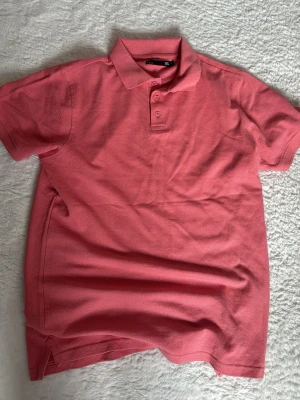 Rosa pikétröja från 157 - Säljer en klassisk rosa pikétröja från 157 med krage och knappar framtill. Tröjan är kortärmad och tillverkad i bomull, perfekt för en avslappnad och stilren look. Passar dig som gillar enkel och snygg stil.