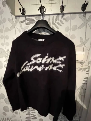 Svart stickad Saint Laurent-tröja - Säljer en svart stickad tröja från Saint Laurent med vit logga i graffiti-stil på bröstet. Tröjan har rund halsringning och långa ärmar. Perfekt för dig som vill ha en trendig och bekväm look med exklusiv känsla. OBS!! REDO ATT GÅ NER I PRIS!