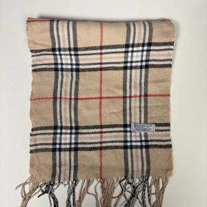 Beige rutig halsduk från Burberrys - Klassisk beige halsduk från Burberrys med ikoniskt rutmönster i svart, vitt och rött. Halsduken har fransar i kanterna och är tillverkad i 100% lammull. Perfekt accessoar för att lyfta din outfit med en tidlös och elegant touch.