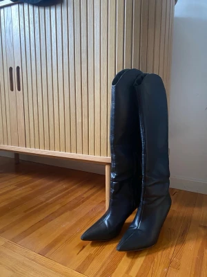 Svarta höga boots med klack - Snygga svarta stövlar i skinnimitation med spetsig tå och smal klack. Stövlarna är knähöga och har en stilren design som passar perfekt till jeans eller kjol. Perfekta för dig som vill ha en trendig och edgy look.