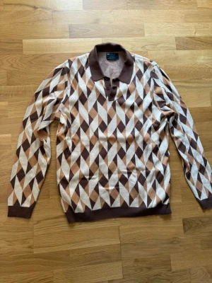 Brun mönstrad tröja från ASOS DESIGN - Säljer en långärmad stickad tröja från ASOS DESIGN med brunt, beige och vitt geometriskt mönster. Tröjan har krage och knappar vid halsen för en retro känsla. Perfekt för dig som gillar unika mönster och snygga detaljer.