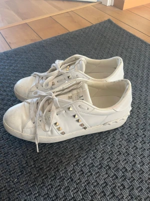 Vita Valentino Garavani sneakers - Snygga vita sneakers från Valentino Garavani med coola guldfärgade nitar på sidorna. Skorna är låga och har klassisk snörning samt en stilren design i läder. Perfekta för dig som vill ha en trendig och exklusiv look.