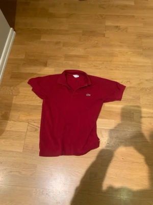 Vinröd pikétröja från Lacoste - Snygg vinröd pikétröja från Lacoste med klassisk krage och korta ärmar. Tröjan har det ikoniska krokodilmärket broderat på bröstet. Perfekt för en stilren och avslappnad look.