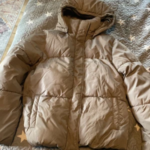 Beige pufferjacka från 157 med huva - Säljer en beige pufferjacka från 157 med huva och dragkedja. Jackan har stora fickor, tryckknappar och är fodrad med mjuk fleece på insidan. Perfekt för kalla dagar och riktigt skön att bära. Passar dig som gillar en enkel och clean stil. Har vissa fläckar men de syns inte på avstånd 😊